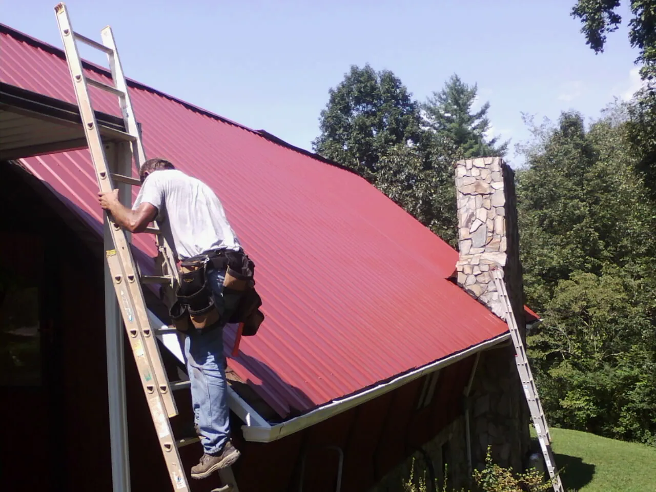 Roofer on a ladder beside a red metal roof and stone chimney — Soffit & Fascia Repair by Roofing Brothers in Nuevo